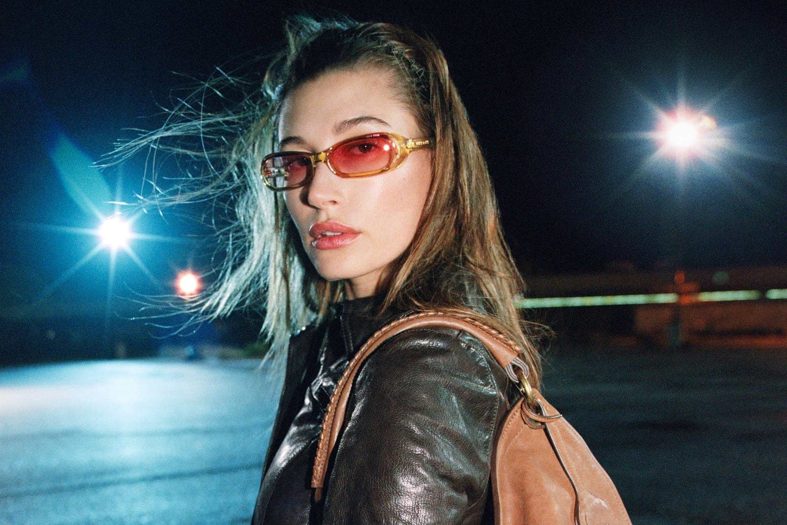 Hailey Bieber photographed in a leather jacket and red sunglasses carrying a tan bag