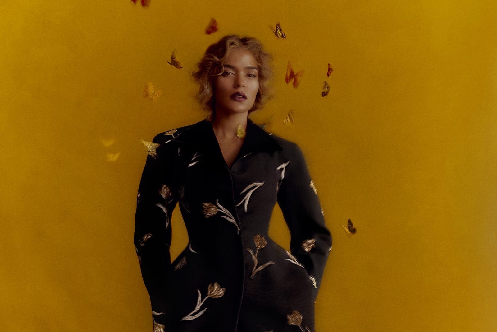Karol G photographed in a floral coat against a yellow background surrounded by butterflies