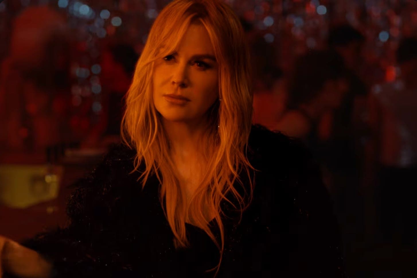 Nicole Kidman in a black coat seated in a dimly lit bar in the She's the Best music video